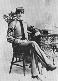 Un fauteuil orné et une table sur laquelle repose un livre. Un homme est assis dans le fauteuil, coiffé d'une casquette à visière de style guerre de Sécession. Sur ses manches, il arbore trois galons pointant vers le bas, surmontés d'un losange, insigne de sergent-chef.