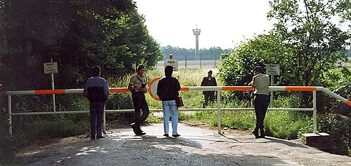 Vue d'une route se terminant par une barrière horizontale rouge et blanche, avec des arbres de chaque côté. Quatre personnes, dont deux en uniforme, se tiennent du côté proche de la barrière. De l'autre côté se trouve un autre homme en uniforme debout dans un champ herbeux. Au fond, une haute clôture métallique et une haute tour de guet avec une cabane octogonale à son sommet.