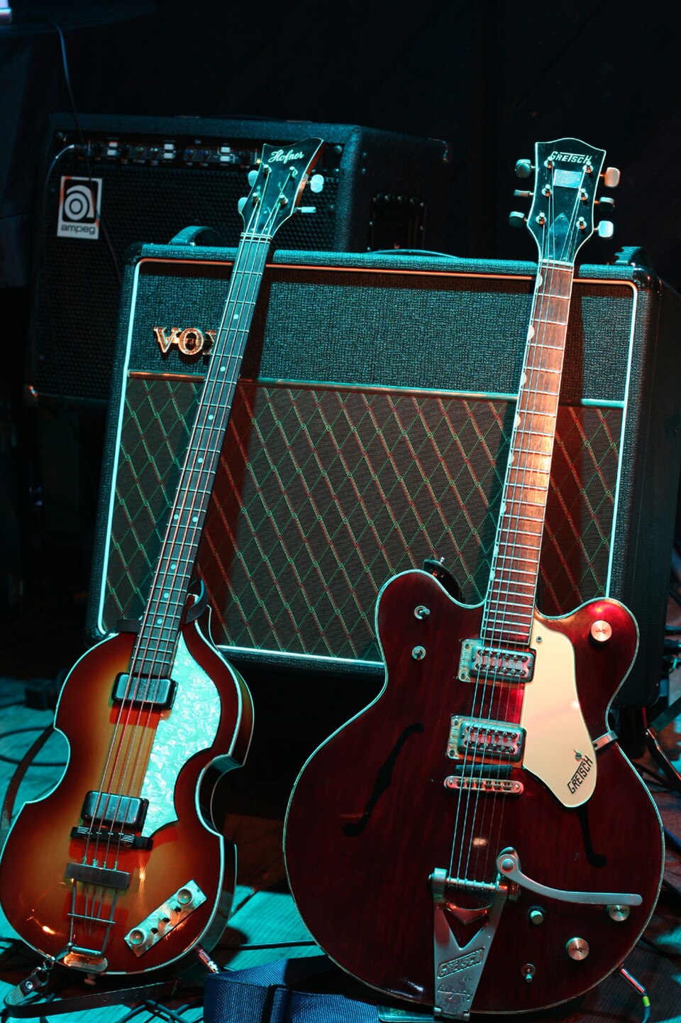 Deux guitares électriques, une basse marron clair en forme de violon et une guitare marron foncé appuyées contre un amplificateur Vox