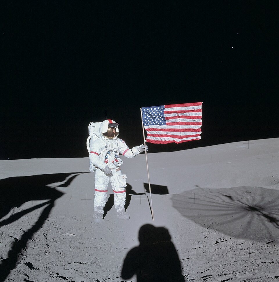 Un astronaute dans une combinaison spatiale Apollo avec des rayures rouges sur les bras, les jambes et le long du casque se tient au milieu de la poussière grise, saisissant le mât d'un drapeau américain