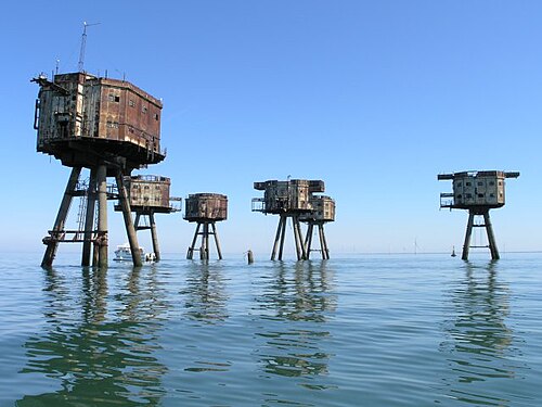 Vue depuis un bateau d'un site contenant six forts maritimes. Les forts ont une forme octogonale, avec des murs en métal rouillé et deux rangées de fenêtres. Chaque fort est soutenu par quatre pieds qui dépassent de la mer à un angle