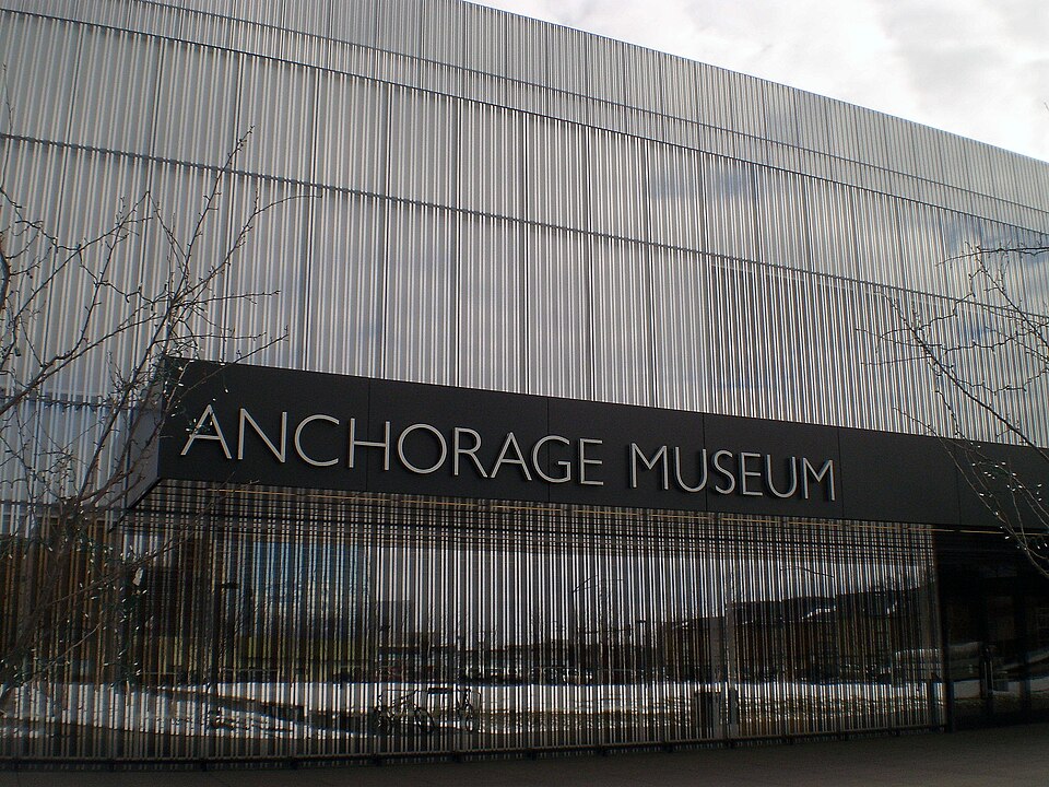 On aperçoit la façade miroitante d'un bâtiment avec une plaque en acier inoxydable portant l'inscription « Anchorage Museum ».