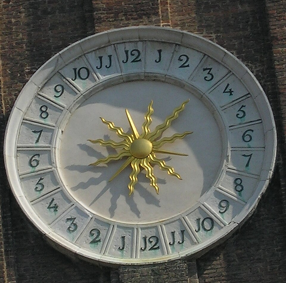 Cadran solaire circulaire décoratif en argile/pierre blanc cassé, orné d'un soleil stylisé doré brillant au centre du cadran 24 heures. Les heures sont graduées de 1 à 12 dans le sens horaire à droite et à gauche, avec des formes en J à l'emplacement des chiffres des unités. Une ombre suggère 15 h en bas à gauche.