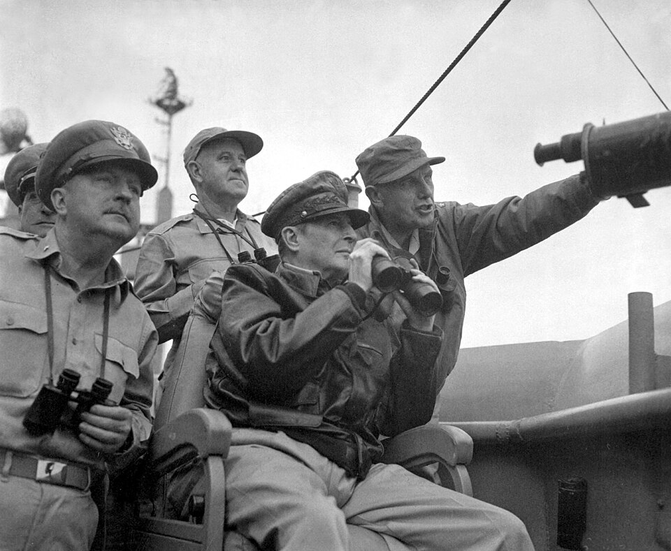 MacArthur est assis, coiffé de son chapeau de maréchal et vêtu d'un blouson aviateur, et tient des jumelles. Quatre autres hommes, également munis de jumelles, se tiennent derrière lui.