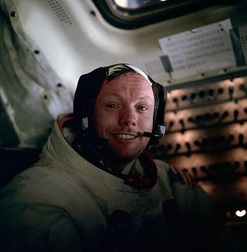 Armstrong sourit dans sa combinaison spatiale sans casque. Il porte un casque et ses yeux semblent légèrement larmoyants.