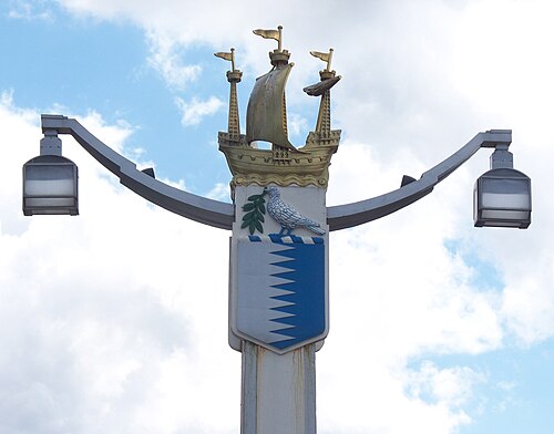 Poteau portant deux lampes à l'extrémité de deux bras. Le sommet du poteau est orné d'un galion ; sous le bateau se trouve un écu composé d'un drapeau blanc à gauche et d'un drapeau bleu à droite, séparés par une ligne brisée, surmonté d'un oiseau blanc portant un rameau d'olivier.