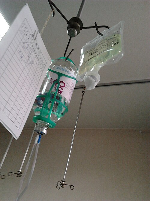 Photographie de deux poches de solution intraveineuse (contenant respectivement du glucose et de la lévofloxacine) et d'une feuille de papier suspendue à un poteau