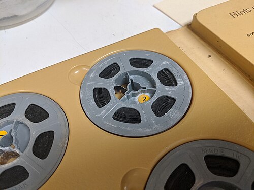 Gros plan d'une bobine de film 8 mm dans un album. La bobine en plastique gris est recouverte d'une substance poudreuse blanche qui s'est répandue sur le film.