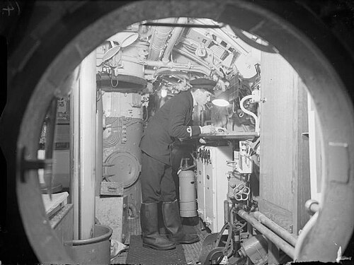 Une photo prise à travers une trappe ronde montre un compartiment rempli de machines. Un homme seul, vêtu d'un uniforme sombre, est penché sur une table, éclairée par une lampe de bureau.