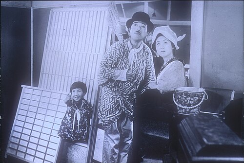 Une jeune fille, un homme et une femme se tiennent devant une maison, tous regardant vers le ciel. La fille, à gauche, sourit et pointe vers le ciel. L'homme porte un chapeau melon et tient un petit balai sur son épaule ; la femme porte un foulard autour de la tête. Ils sont entourés d'objets domestiques comme s'ils venaient d'entrer ou de sortir de la maison.
