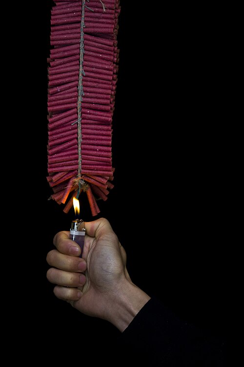 Une guirlande de pétards rouges. Un jeune homme tient une flamme pour l'allumer.