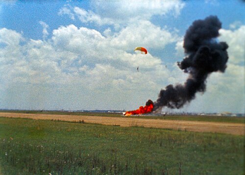 Une photo indistincte d'une traînée de fumée s'élevant d'une zone d'incendie orange dans un champ récemment récolté. Un parachute blanc et orange recouvre une silhouette humaine au-dessus et à droite de l'incendie.