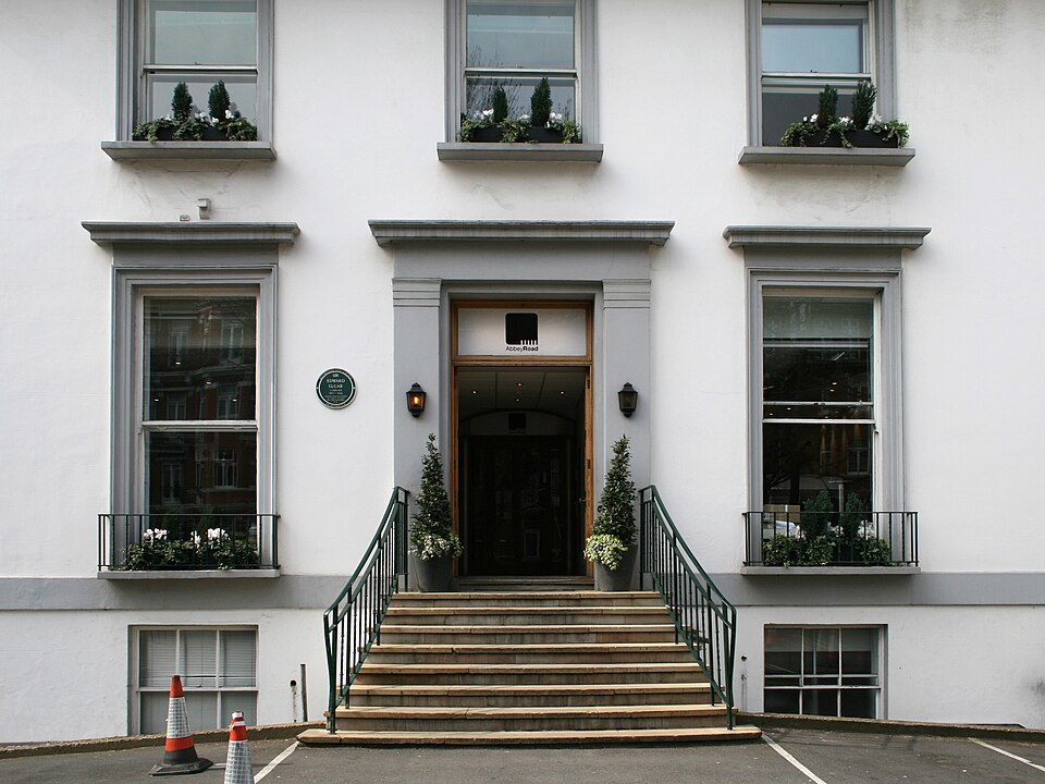 Un escalier en pierre mène d'un parking asphalté à l'entrée principale d'un bâtiment blanc de deux étages. Le rez-de-chaussée possède deux fenêtres à guillotine, le premier étage trois, plus courtes. Deux autres fenêtres sont visibles au sous-sol. Les ornements en pierre autour des portes et des fenêtres sont peints en gris.