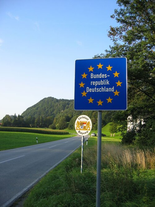 Deux panneaux routiers sur le côté d'un tronçon ouvert d'une autoroute à deux voies. Un poste frontière interne de l'espace Schengen signalé uniquement par un panneau bleu indiquant le pays d'entrée. Le panneau blanc plus petit annonce l'entrée dans l'État de Bavière.