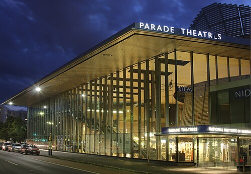 Une vue extérieure du bâtiment de nuit. Il est bien éclairé et possède des panneaux de verre sur presque toute la longueur. Sur le côté droit se trouvent le nom de l'école d'art dramatique ainsi que les lettres de Parade Theatres. Un escalier intérieur est visible sur le côté gauche et une structure en bois arrondie à droite.