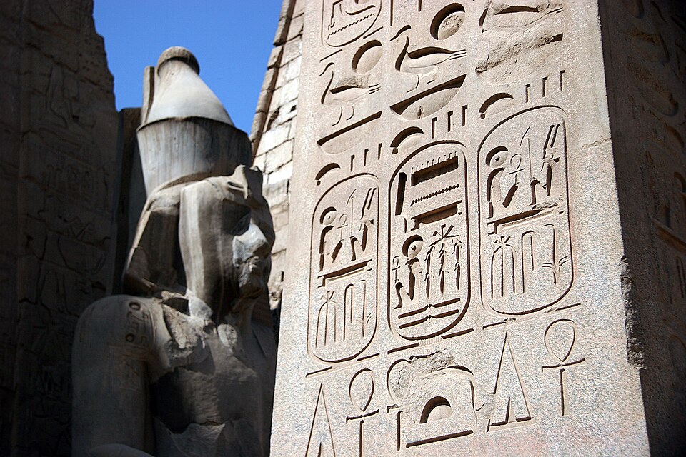 Des hiéroglyphes inscrits recouvrent un obélisque au premier plan. Une statue de pierre se dresse à l'arrière-plan.