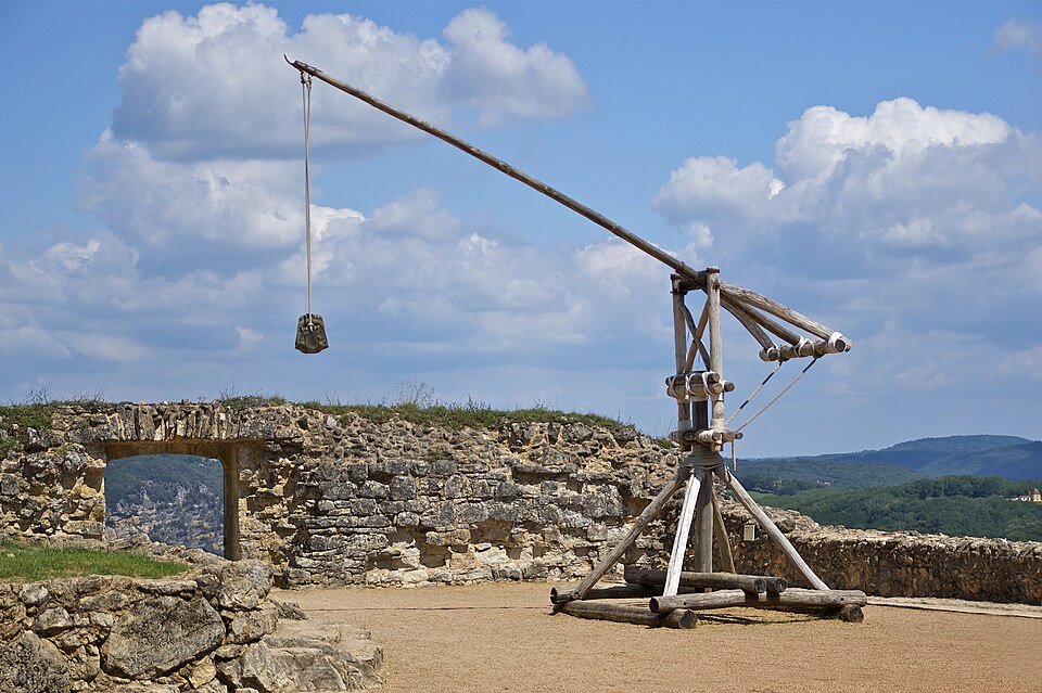 Photo d'un trébuchet médiéval reconstitué sur une plateforme en pierre, avec un ciel nuageux en arrière-plan.