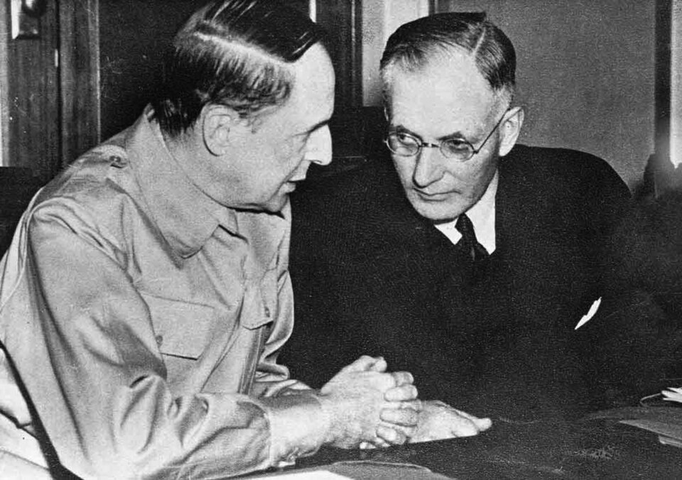 Deux hommes sont assis côte à côte à une table et discutent. L'un porte un costume, l'autre un uniforme militaire.