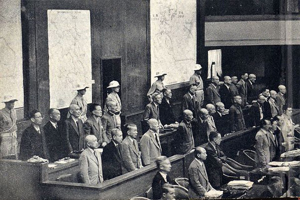 Trois rangées de bancs, derrière lesquelles se tiennent une douzaine d'hommes environ. Derrière eux se tiennent cinq hommes en uniforme.
