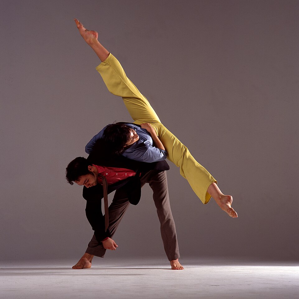 Danse moderne – une danseuse exécute un grand écart facial en équilibre sur le dos de son partenaire.