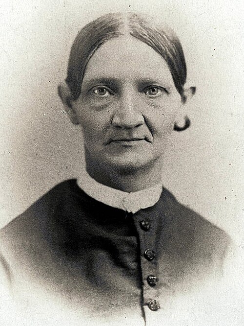 Photographie sépia ancienne de Lucy Smith Millikin, prise entre 1850 et 1900. La femme âgée a les cheveux tirés en chignon, des oreilles proéminentes et des épaules fines. Elle est vêtue d'une robe sombre avec un col blanc.