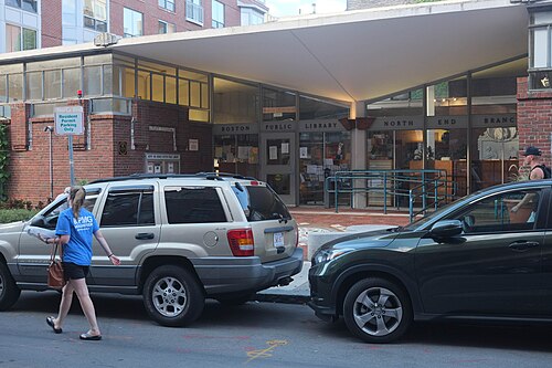 L'extérieur de la succursale North End de plain-pied du système de bibliothèque publique de Boston. Deux voitures sont garées devant et une personne portant un t-shirt bleu et un short noir marche dans la rue. Le bâtiment est principalement en brique, mais il y a une section de portes et de fenêtres pour l'entrée.