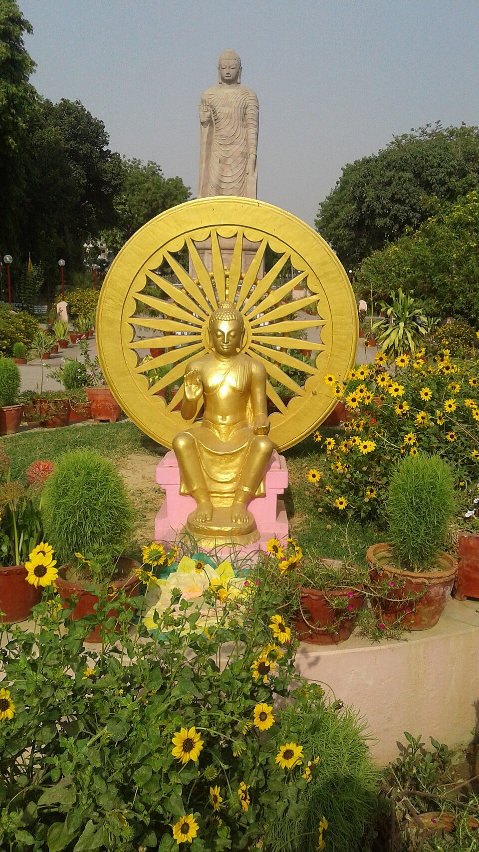 Statue du Bouddha assis, avec une statue du Bouddha debout en arrière-plan, sur le site du temple et du monastère thaïlandais de Sarnath.