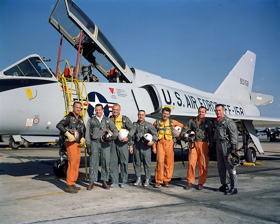 Les astronautes posent devant un avion à réaction bleu-gris clair à ailes delta, tenant leur casque de vol sous le bras. Les trois aviateurs de la Navy portent des combinaisons de vol orange, celles de l'Air Force et des Marines sont vertes.