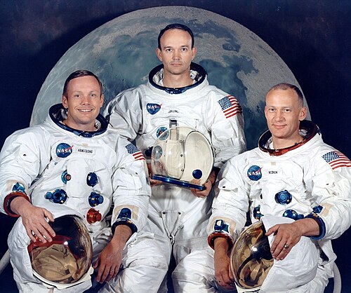 Trois astronautes en combinaison spatiale blanche. Ils tiennent leur casque. Ils ont tous la peau claire. Armstrong sourit largement et porte les cheveux séparés sur la droite. Collins a les cheveux foncés et a l'air le plus sérieux. Les cheveux d'Aldrin sont très courts. Derrière eux se trouve une grande photo de la Lune.
