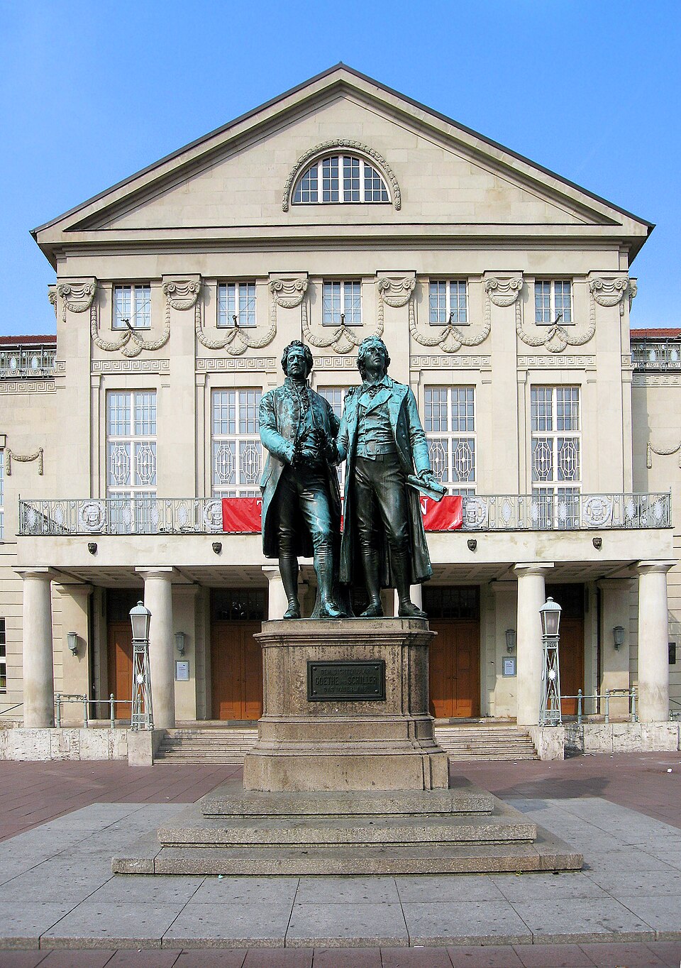 Photographie d'une grande statue en bronze représentant deux hommes côte à côte, de face. La statue repose sur un piédestal en pierre portant une plaque où l'on peut lire « Dem Dichterpaar/Goethe und Schiller/das Vaterland ». Derrière le monument se dresse un grand bâtiment de trois étages à la façade en pierre richement ornée.