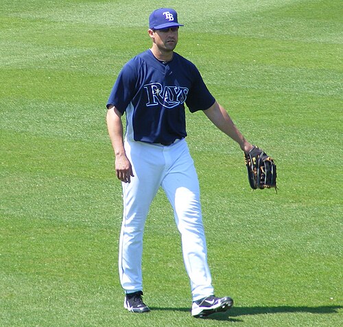 Un homme debout sur une surface herbeuse portant un uniforme de baseball avec une chemise bleu foncé et un pantalon blanc