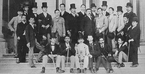 Photographie d'un grand groupe d'hommes sur des marches. Certains sont assis, d'autres sont debout ; plusieurs portent des chapeaux haut-de-forme.