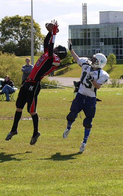 Deux joueurs de football, un receveur offensif et un cornerback défensif, tentent tous deux de récupérer un ballon lancé. Le cornerback se trouve devant le receveur avec le ballon presque en sécurité dans ses mains.