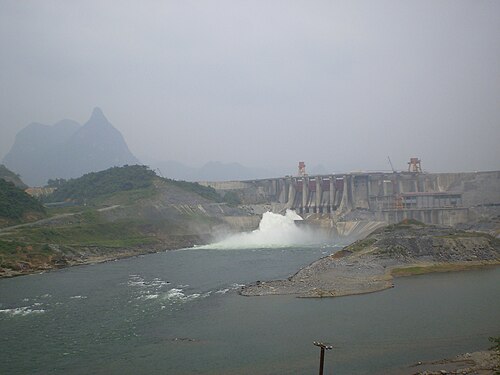 Au loin, un barrage gris déverse de l'eau depuis son centre. Les montagnes sont en arrière-plan.