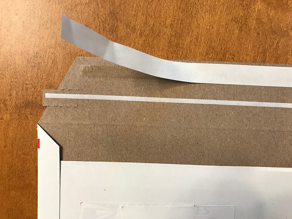 Enveloppe d'expédition en carton avec adhésif PSA, film protecteur et bande d'arrachage.