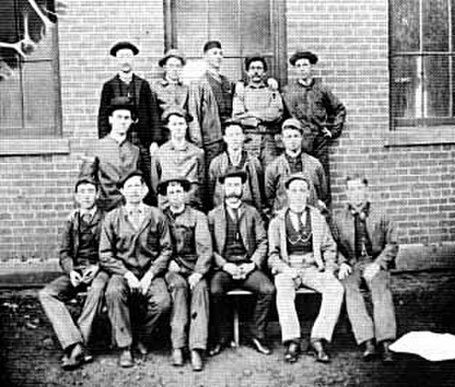 Photo de groupe de quinze hommes ; cinq dans la rangée du haut, quatre au milieu et six au premier plan, posant devant un bâtiment en briques