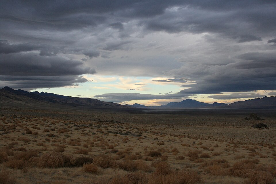Paysage d'une longue vallée aride. Le ciel est partiellement nuageux, mais des éclaircies bleues percent par endroits. C'est un véritable panorama de la beauté naturelle du Nevada.