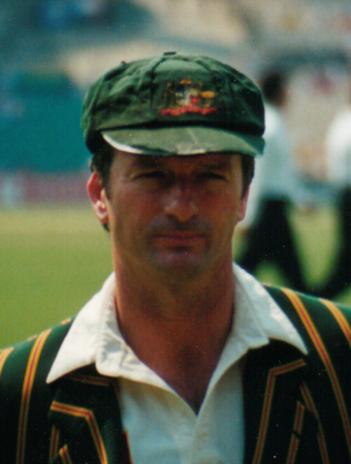 Homme d'âge moyen d'environ 30 ans portant une casquette verte ample avec les armoiries australiennes, un blazer australien vert à rayures jaunes et une chemise de cricket couleur crème. Il est rasé de près et a les cheveux bruns.