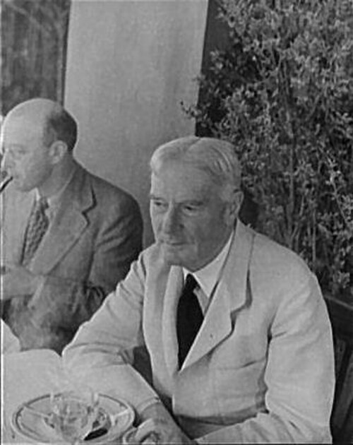 photographie extérieure d'un homme âgé assis à une table ; il a les cheveux blancs et est rasé de près