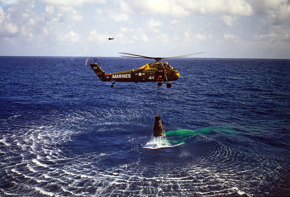 Un hélicoptère vert plane au-dessus de l'eau, avec la capsule Mercury suspendue en dessous. L'hélicoptère porte l'inscription « Marines » et le numéro « 44 »