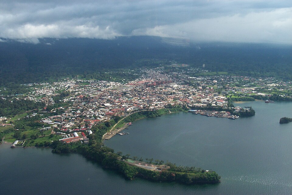 Au loin, on voit des montagnes couvertes de nuages, tandis qu'à l'avant de l'image se trouvent des maisons menant au port qui possède deux jetées menant à la mer.