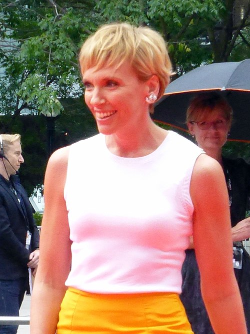 La femme de 42 ans est photographiée à mi-corps. Elle porte un haut blanc sur une jupe jaune. Elle sourit et regarde vers la gauche avec ses cheveux courts, châtain clair à châtain clair. Derrière elle, à notre gauche, se trouve un homme portant des écouteurs et un microphone, tandis qu'à droite, une femme tient un parapluie.