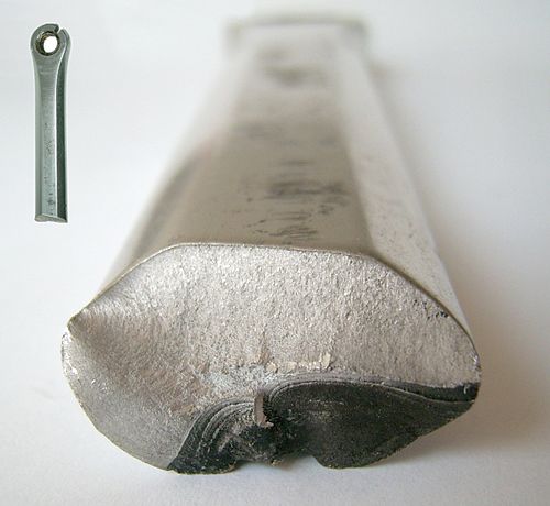 Cylindre métallique de forme grossièrement ovoïde, vu de face. La partie inférieure droite de la surface terminale du métal est sombre et légèrement déformée, tandis que le reste est d'une couleur beaucoup plus claire et n'est pas déformé.