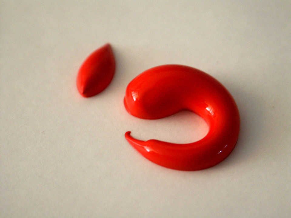 Une tache de peinture acrylique rouge en forme de limace brille sur une surface blanche, avec une petite tache en haut à gauche.
