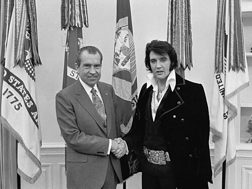 Presley, au physique de mouton, porte une longue veste en velours et une boucle géante comme celle d'une ceinture de championnat de boxe. Il serre la main d'un homme chauve portant un costume et une cravate. Ils font face à la caméra et sourient. Cinq drapeaux sont suspendus à des poteaux juste derrière eux.