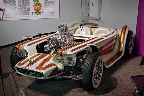 Une voiture blanche à rayures marron, avec des roues ouvertes et un toit transparent surmontant deux sièges, et un moteur chromé exposé avec un ventilateur. Beatnik Bandit II et quelques autres voitures de Roth sont également exposées dans ce musée.