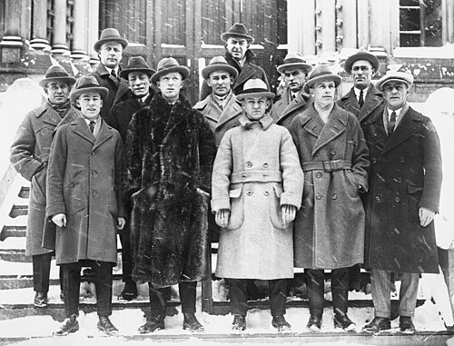 Douze hommes posent sur les marches devant un immeuble. Ils portent des costumes, de longues vestes et des chapeaux.