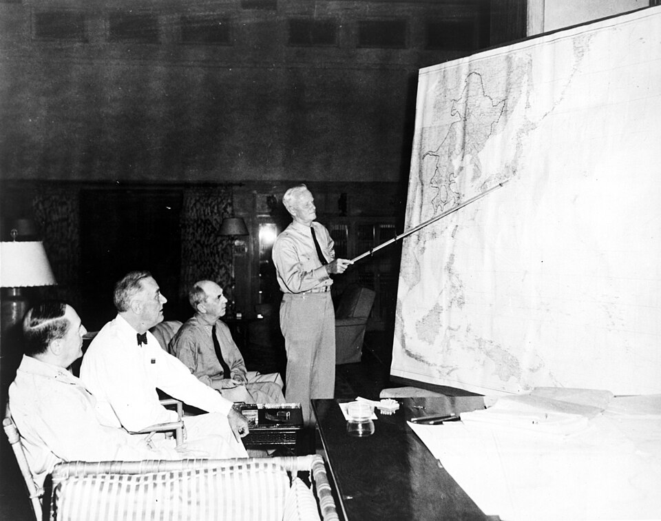 Trois hommes sont assis dans des fauteuils. L'un d'eux est debout, tenant un long bâton et désignant l'emplacement du Japon sur une carte murale du Pacifique.