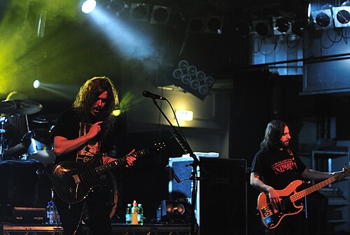 Deux hommes aux cheveux longs sont sur une scène. L'un joue d'une guitare brillante et l'autre d'une guitare basse avec une finition en bois