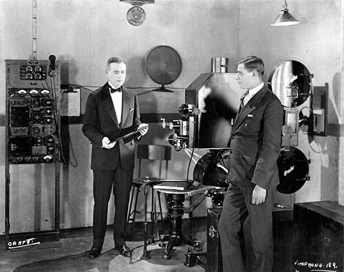 Deux hommes en costume se tiennent dans un studio équipé d'un grand projecteur de cinéma et d'autres équipements électriques. L'homme de gauche tient un grand disque de phonographe.